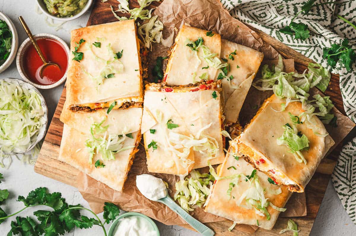 Overhead view of baked Crunchwrap Casserole cut into squares on a wooden board, topped with shredded lettuce and cilantro, surrounded by bowls of guacamole, sour cream, and hot sauce.