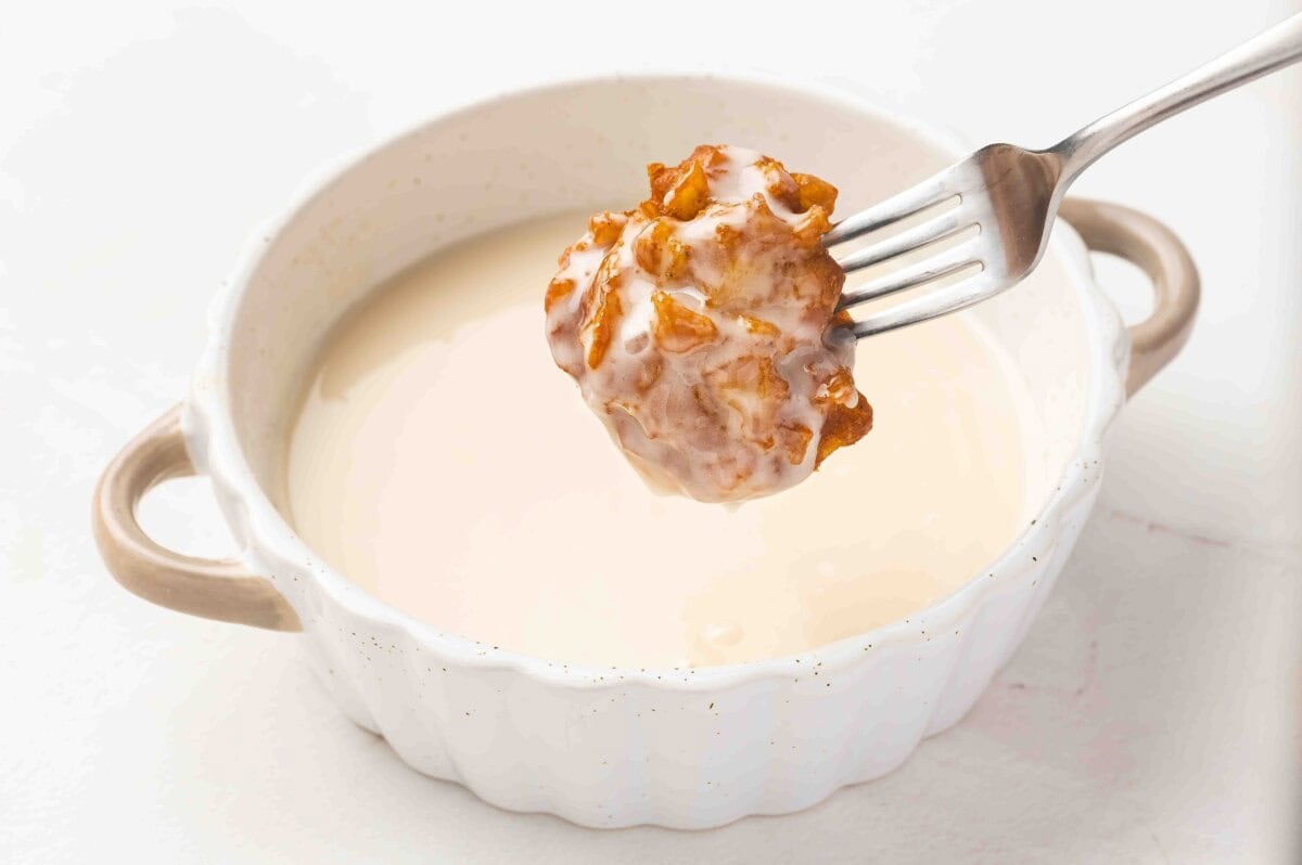 A golden apple fritter held with a fork being dipped into a bowl of vanilla glaze.