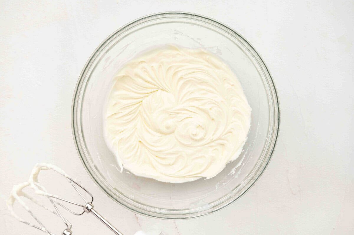 A glass bowl filled with whipped mascarpone cream, smooth and fluffy, with a hand mixer resting nearby.
