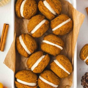 A white dish lined with parchment paper filled with pumpkin whoopie pies sandwiched with cream cheese frosting, surrounded by cinnamon sticks and fall decorations.