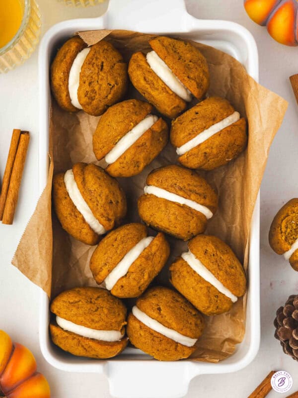 A white dish lined with parchment paper filled with pumpkin whoopie pies sandwiched with cream cheese frosting, surrounded by cinnamon sticks and fall decorations.