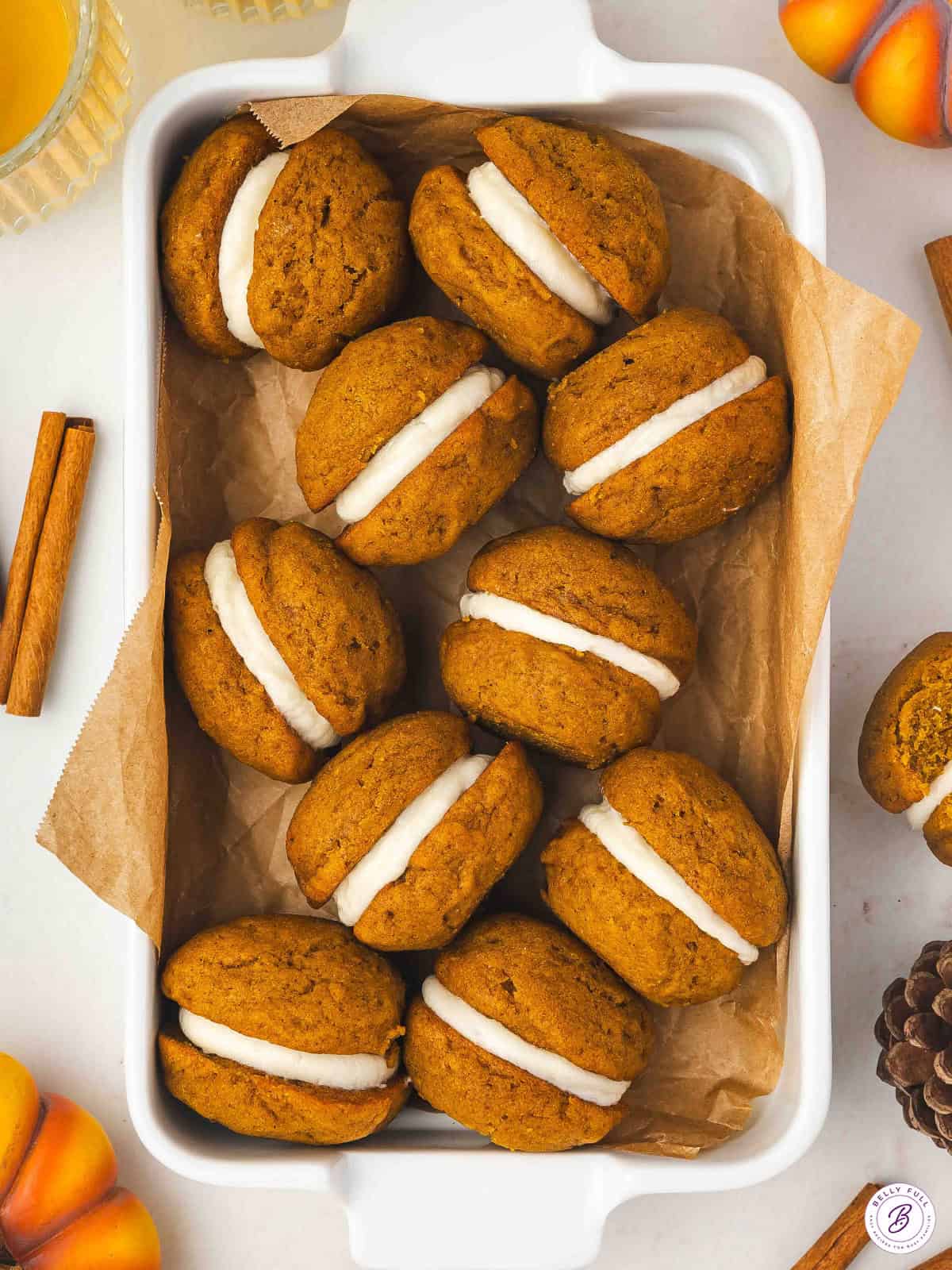 A white dish lined with parchment paper filled with pumpkin whoopie pies sandwiched with cream cheese frosting, surrounded by cinnamon sticks and fall decorations.