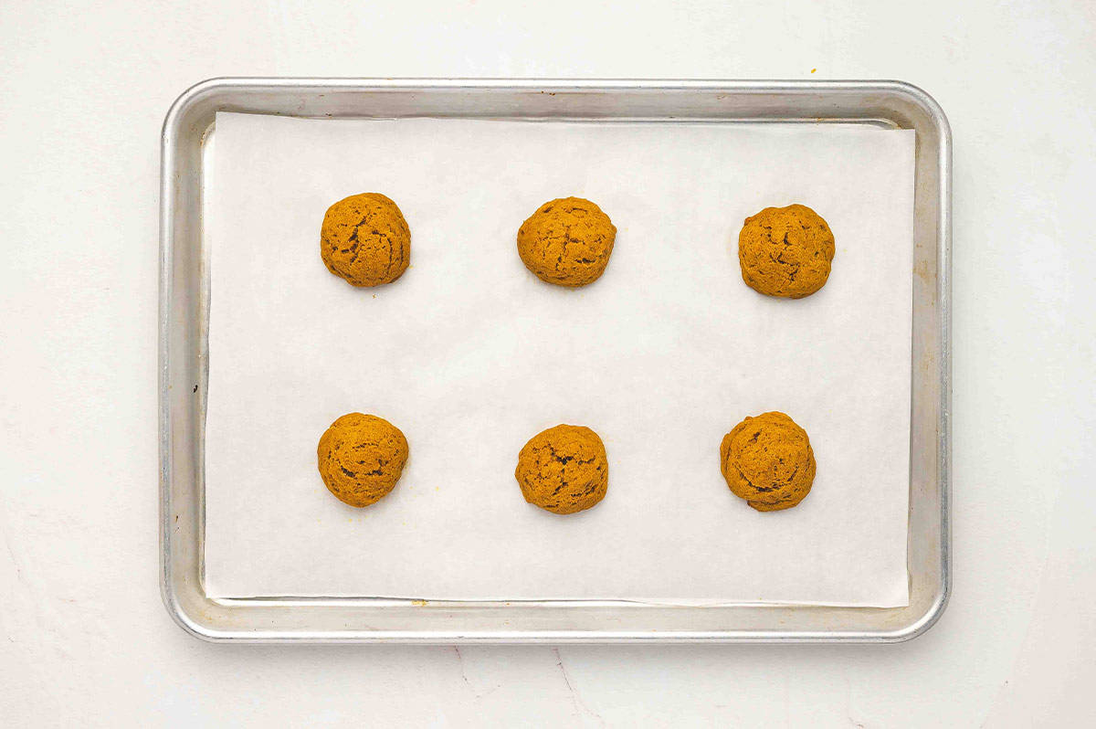 A baking sheet lined with parchment paper holding six baked pumpkin cake rounds cooling after baking.