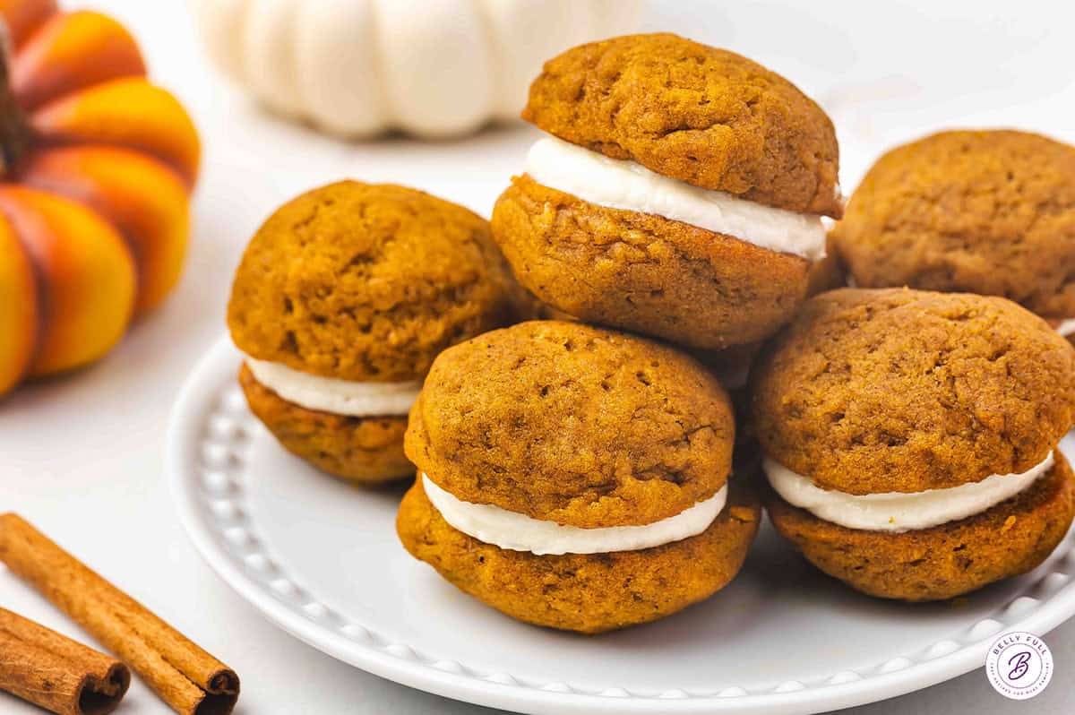A white plate with pumpkin whoopie pies stacked and ready to serve, surrounded by pumpkins and cinnamon sticks.