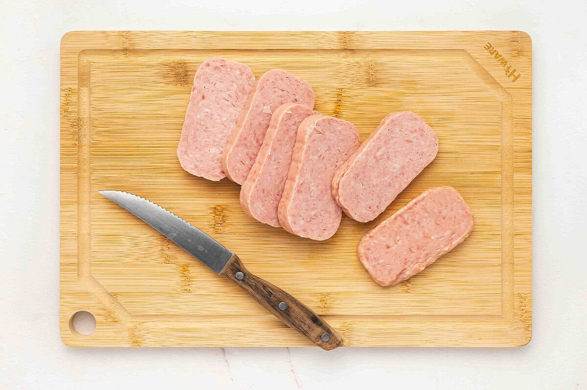Six slices of raw Spam on a wooden cutting board with a small serrated knife.