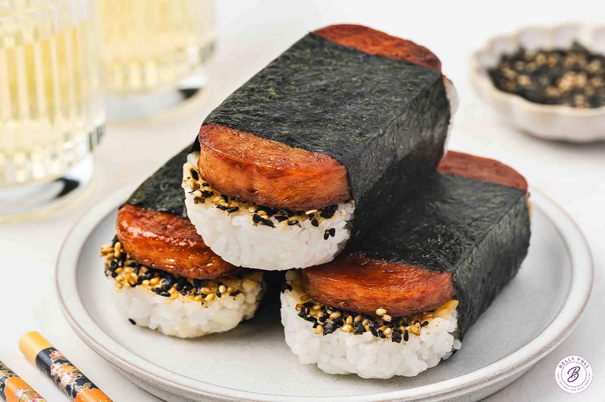 Three Spam Musubi pieces lined up on a white rectangular plate with rice, furikake, glazed spam, and nori wrap, with chopsticks nearby