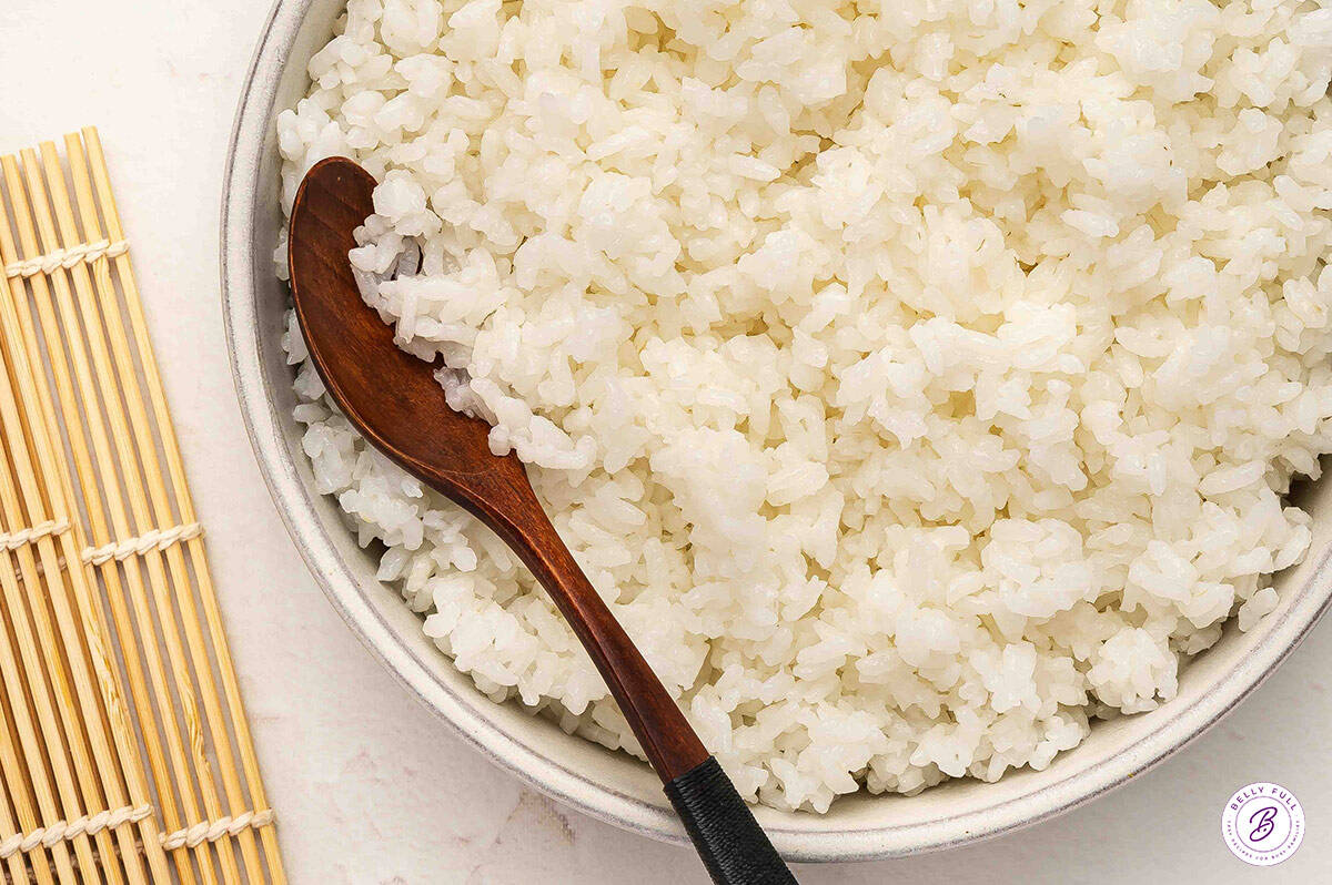 Close-up of glossy sushi rice in a ceramic bowl with a wooden spoon, next to a bamboo sushi mat.