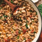 Overhead view of Tuscan white bean and sausage skillet in a pan, with a wooden spoon lifting a serving of beans, sausage, spinach, and sun-dried tomatoes.