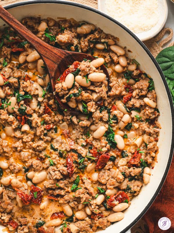 Overhead view of Tuscan white bean and sausage skillet in a pan, with a wooden spoon lifting a serving of beans, sausage, spinach, and sun-dried tomatoes.