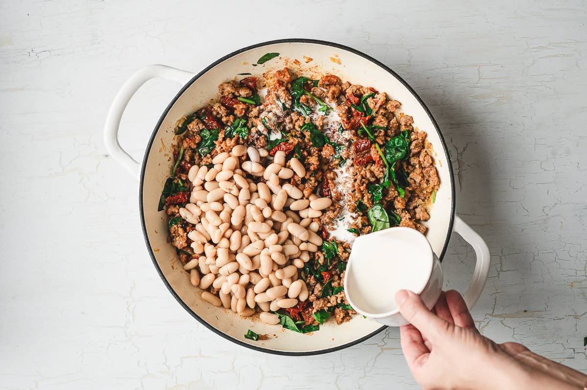 Hand pouring cream into a skillet filled with cooked sausage, spinach, sun-dried tomatoes, and cannellini beans.