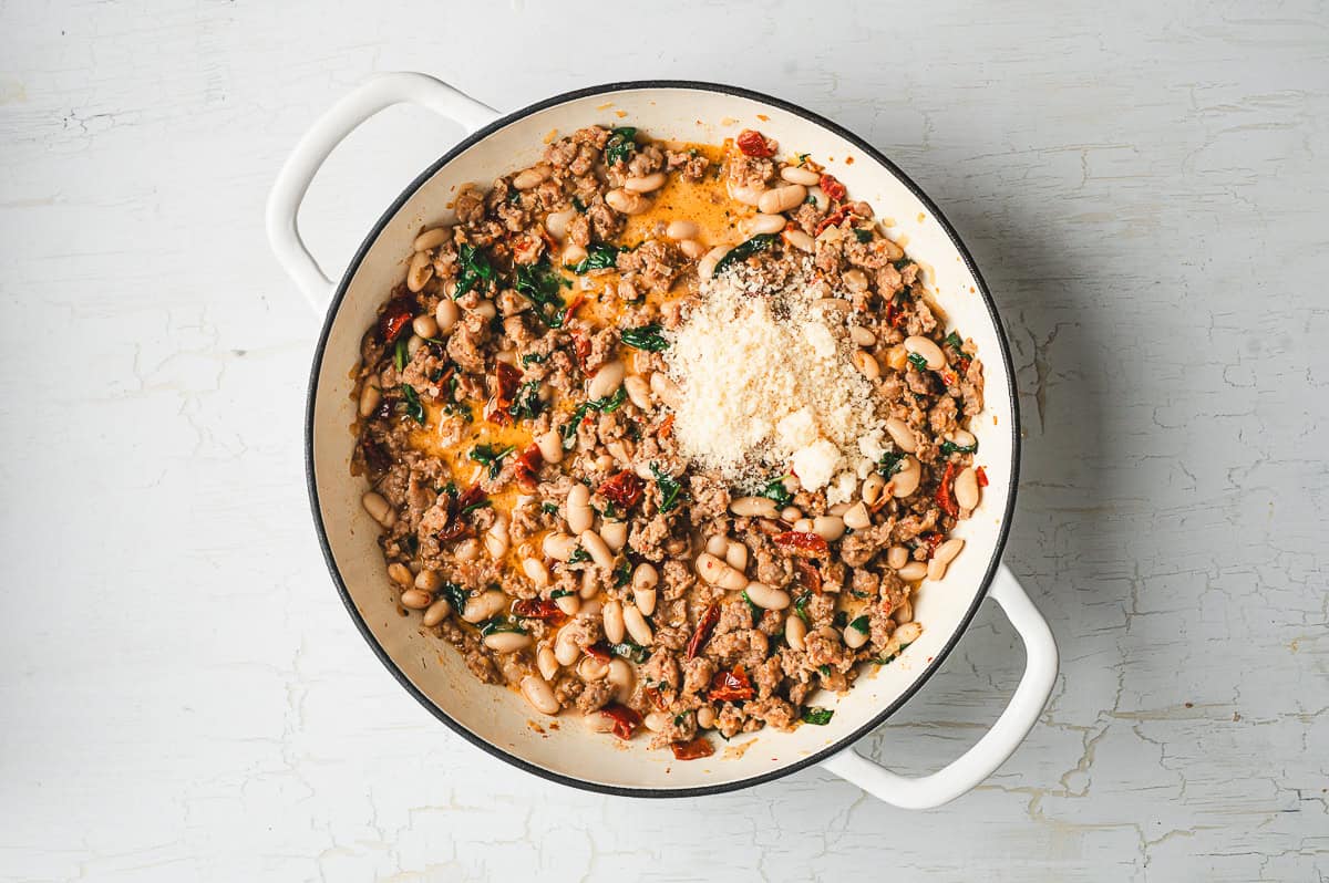 Cooked sausage and beans mixture in a skillet topped with a mound of freshly grated Parmesan cheese before stirring.