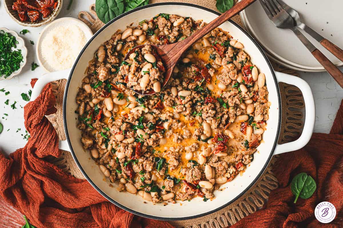 Large white skillet filled with Tuscan white bean and sausage mixture, garnished with parsley, surrounded by bread, cheese, and plates on the table.