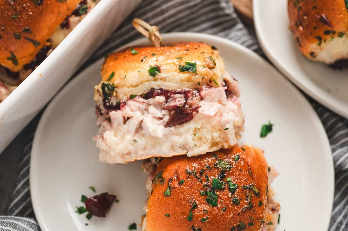 Single cranberry turkey slider on a small plate showing gooey melted cheese, cranberry sauce, and golden bun sprinkled with herbs.
