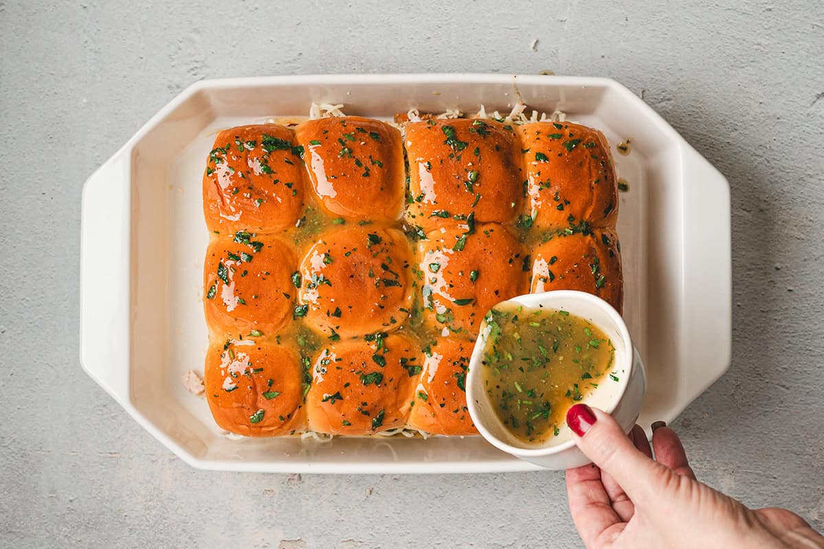 Top halves of slider rolls placed on and coated with warm buttery honey-mustard glaze using a small cup.