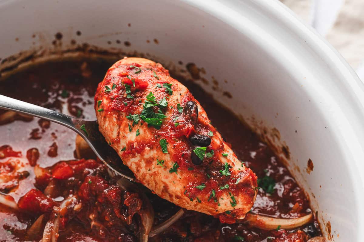A cooked chicken breast being lifted from the slow cooker, coated in tomato balsamic sauce and garnished with parsley.