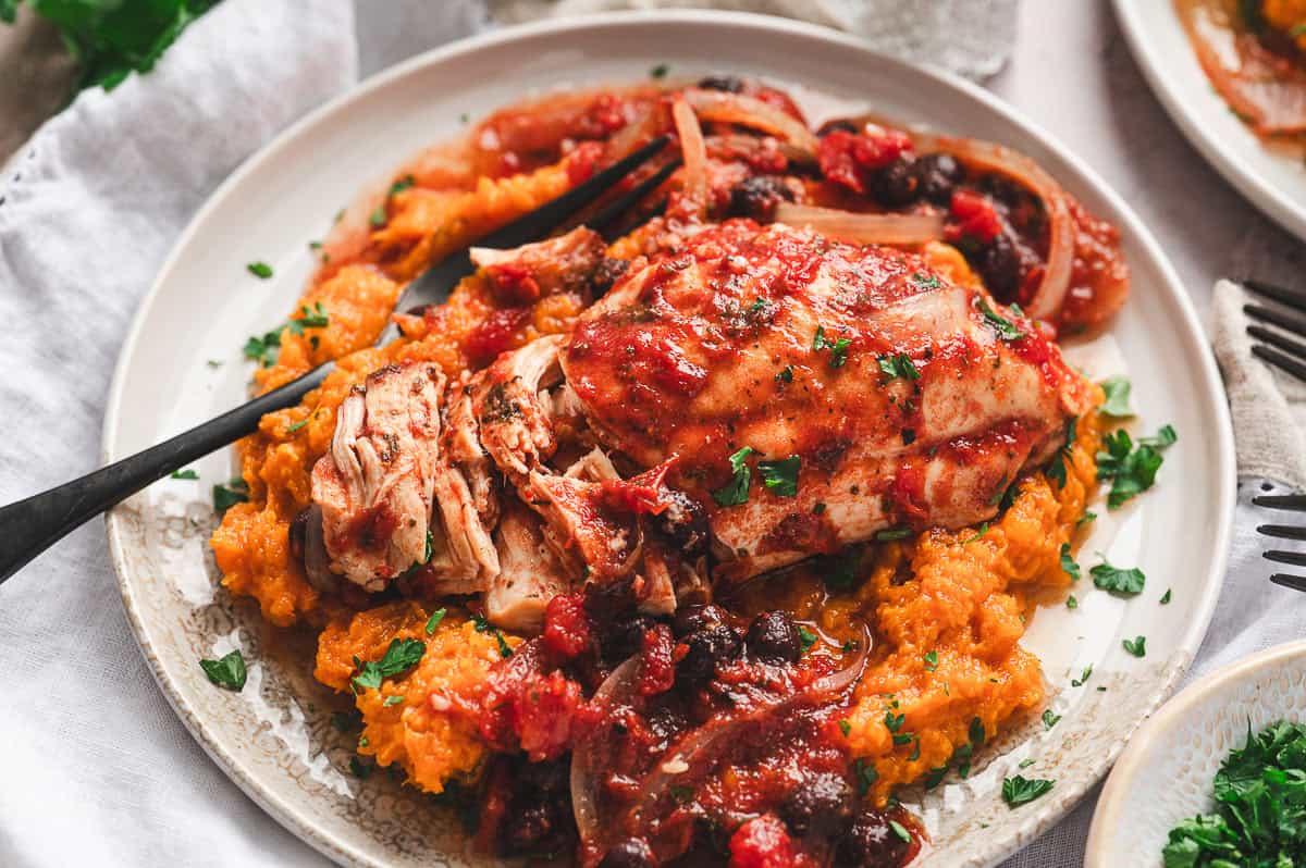 Slow cooker tomato balsamic chicken served over mashed sweet potatoes on a plate with fresh parsley.