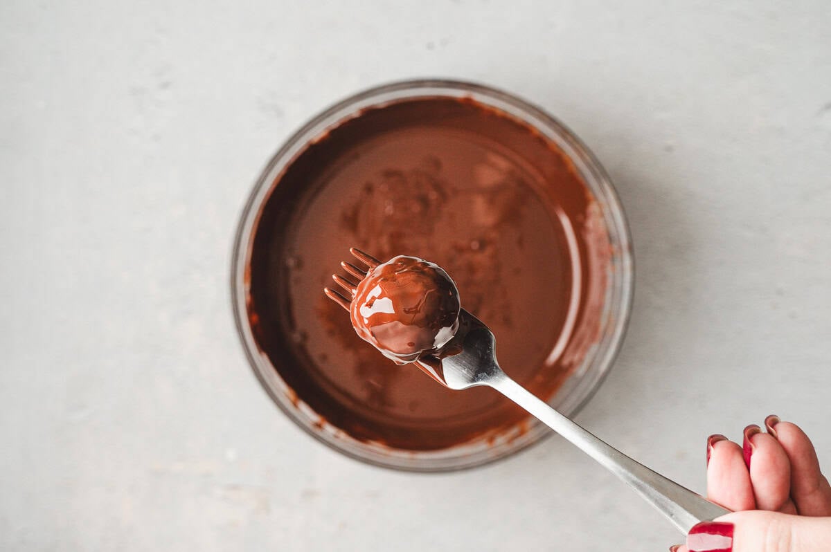 A fork holding one kataifi truffle being dipped into glossy melted dark chocolate.