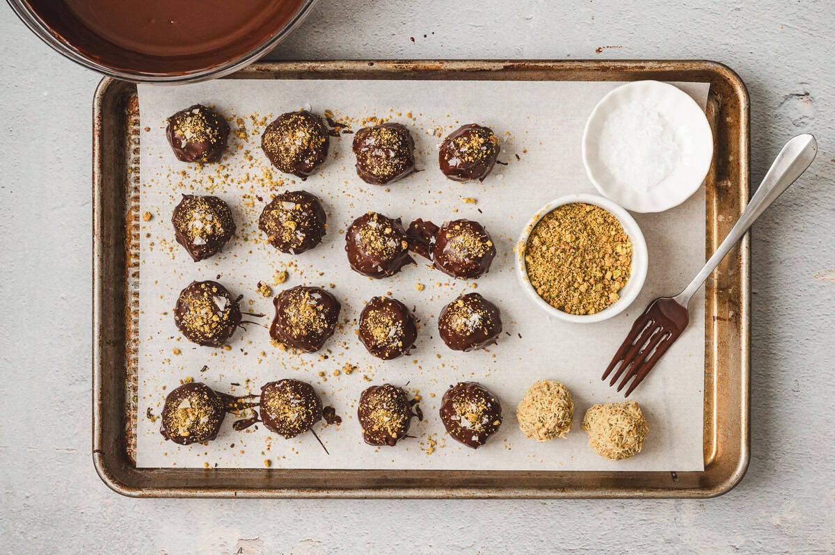 Chocolate-coated truffles on a parchment-lined tray, sprinkled with crushed pistachios and sea salt, with dipping bowl and garnish dishes beside them.