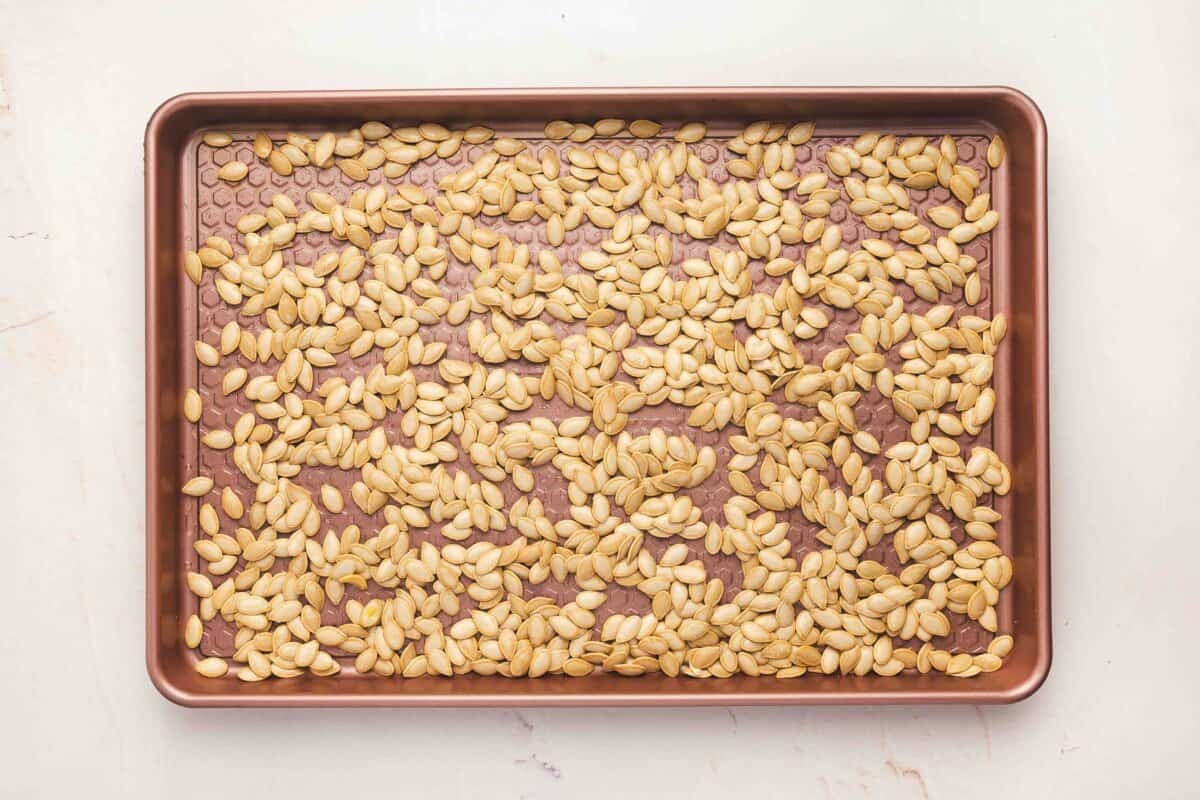 Seasoned pumpkin seeds spread in a single layer on a rimmed baking sheet, ready to roast
