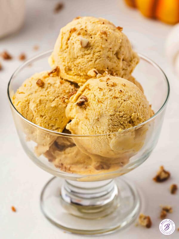 Three scoops of no-churn pumpkin pie ice cream in a glass dessert bowl topped with chopped candied pecans.