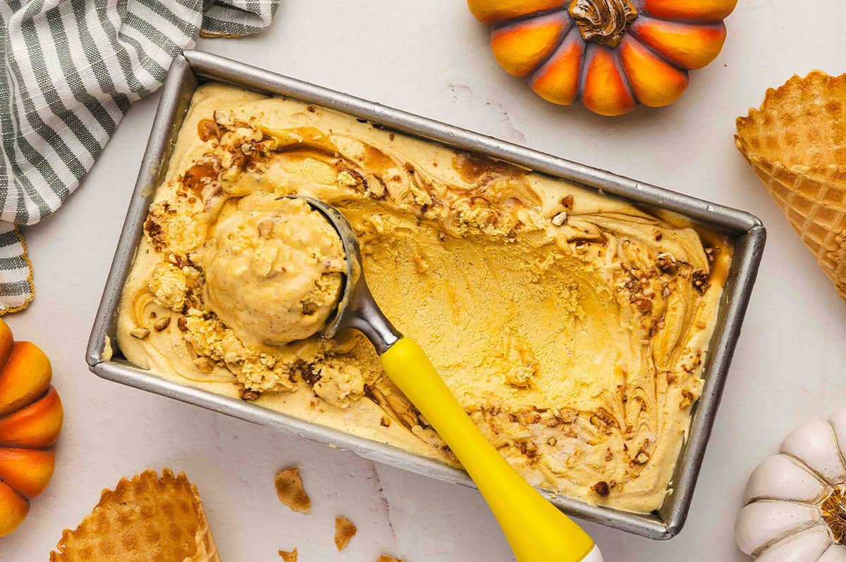 Partially scooped loaf pan of frozen pumpkin pie ice cream with a yellow-handled ice cream scoop, waffle cones and pumpkins around it.