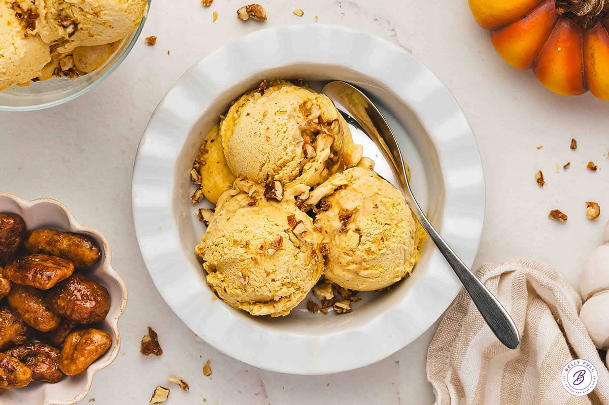 Three scoops of pumpkin pie ice cream in a white bowl, sprinkled with chopped pecans and served with a spoon, surrounded by fall decorations.