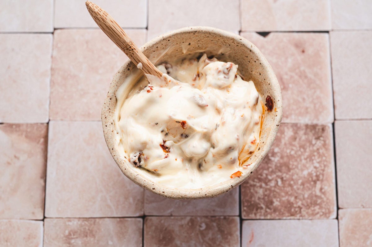 Small bowl of mayonnaise mixed with chopped sun-dried tomatoes, with a wooden spoon inside.
