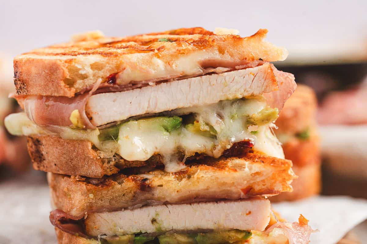 Stacked slice of panini showing the layers of turkey, prosciutto, avocado, and melted Swiss cheese oozing out.