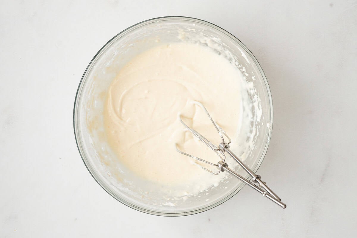 A glass bowl filled with smooth cream cheese filling and electric mixer beaters resting inside.