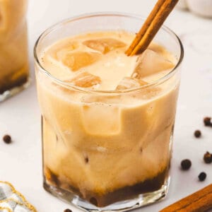 A creamy pumpkin pie White Russian served in a lowball glass filled with ice and topped with a cinnamon stick, with warm fall spices and pumpkins in the background.