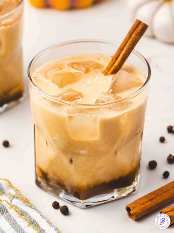 A creamy pumpkin pie White Russian served in a lowball glass filled with ice and topped with a cinnamon stick, with warm fall spices and pumpkins in the background.