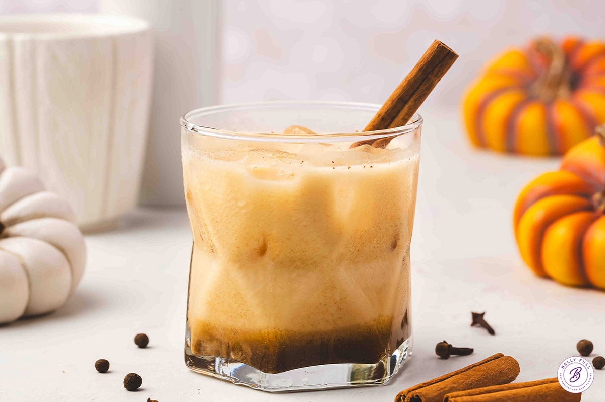 Close-up of a finished pumpkin pie White Russian with a cinnamon stick garnish, surrounded by cinnamon sticks and small pumpkins for a cozy fall presentation.