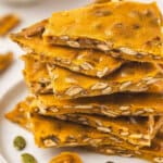 Stack of golden pumpkin seed toffee brittle pieces on a white plate with pumpkin seeds scattered around.