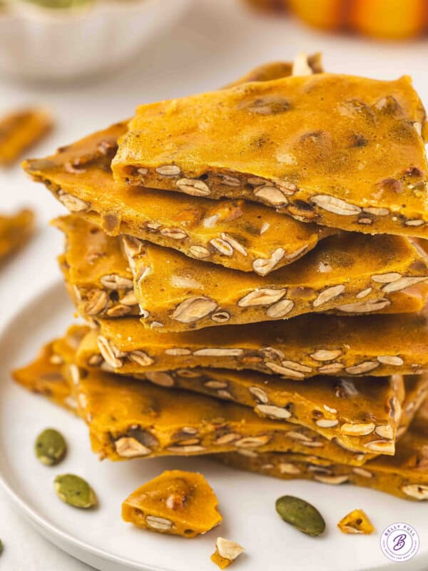 Stack of golden pumpkin seed toffee brittle pieces on a white plate with pumpkin seeds scattered around.