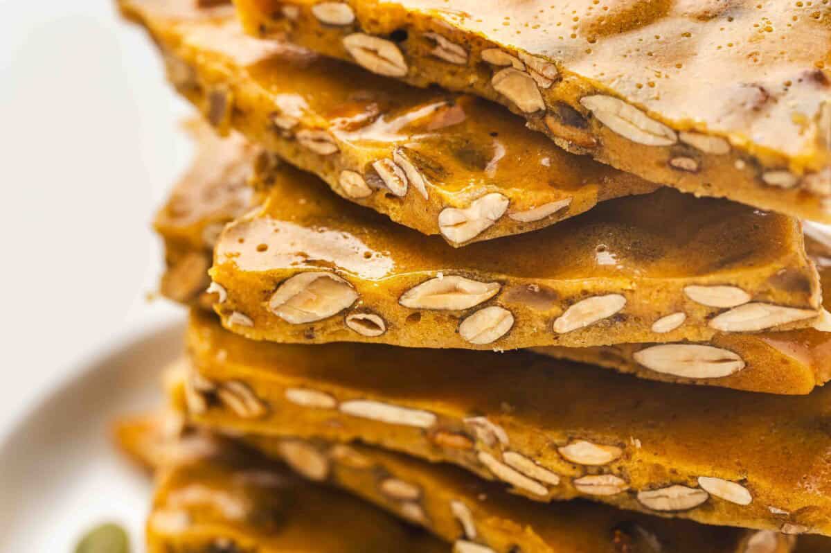 Close-up view of golden pumpkin seed toffee brittle stacked on a white plate, showing its glossy caramel coating and crunchy roasted pumpkin seeds inside.