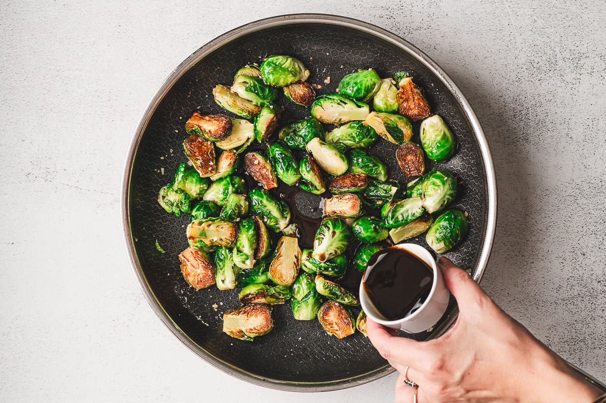Hand pouring a small bowl of molasses mixture into a skillet filled with sautéed Brussels sprouts.