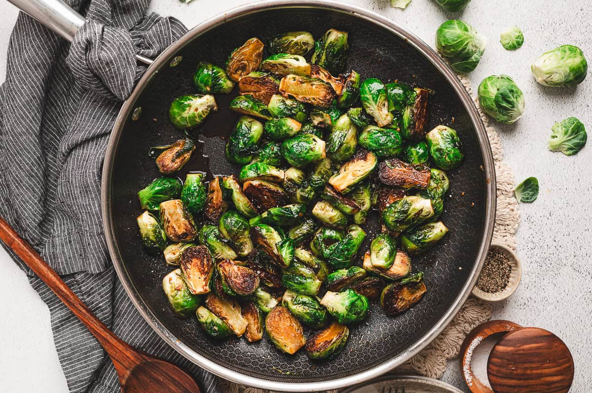 Brussels sprouts in a skillet, coated in the molasses glaze, with a kitchen towel and wooden spoon beside the pan.