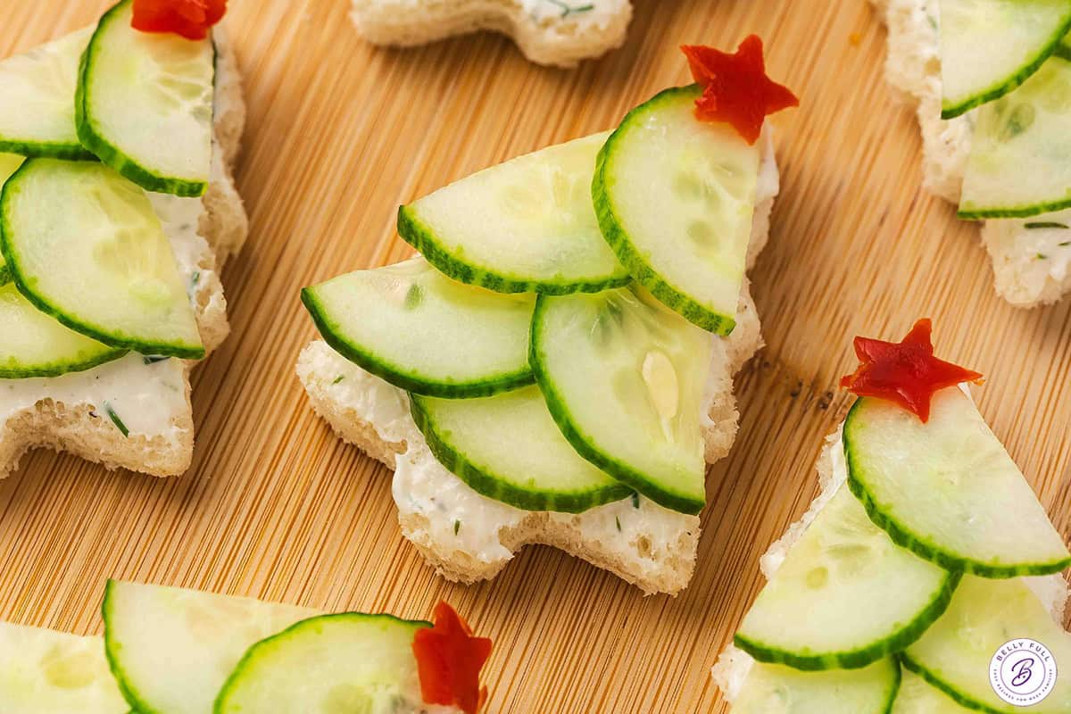 Close-up of festive Christmas tree cucumber sandwiches made with herbed cream cheese, layered cucumber slices, and red bell pepper stars on top.