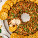 Christmas wreath cheese ball coated in chopped almonds, sun-dried tomatoes, and parsley, arranged in a wreath shape and served with round butter crackers on a platter, with a portion scooped out.