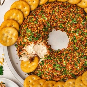 Christmas wreath cheese ball coated in chopped almonds, sun-dried tomatoes, and parsley, arranged in a wreath shape and served with round butter crackers on a platter, with a portion scooped out.