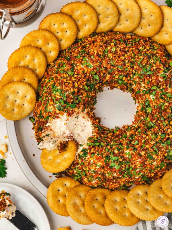Christmas wreath cheese ball coated in chopped almonds, sun-dried tomatoes, and parsley, arranged in a wreath shape and served with round butter crackers on a platter, with a portion scooped out.