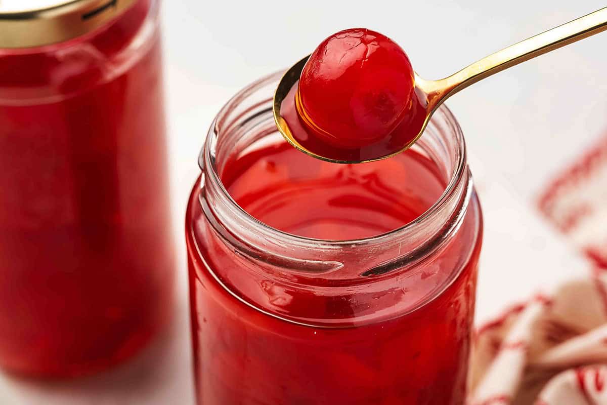 A spoon lifting a vodka soaked maraschino cherry from a jar filled with bright red liquid.