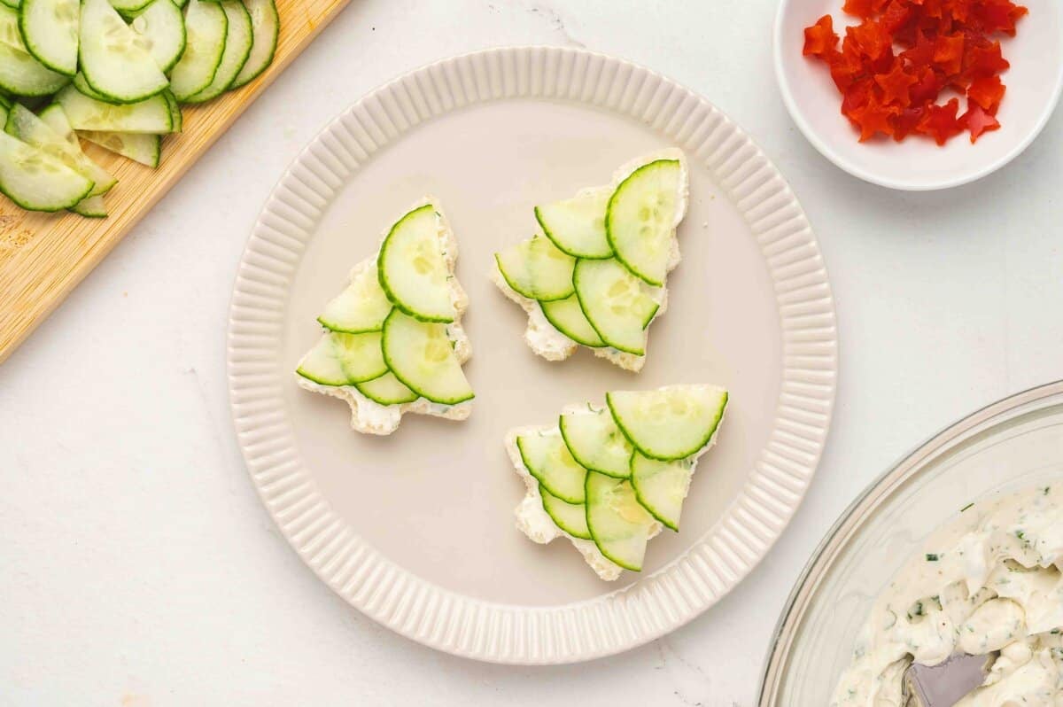 Partially assembled Christmas tree sandwiches on a plate with cucumber slices and bell pepper pieces nearby.
