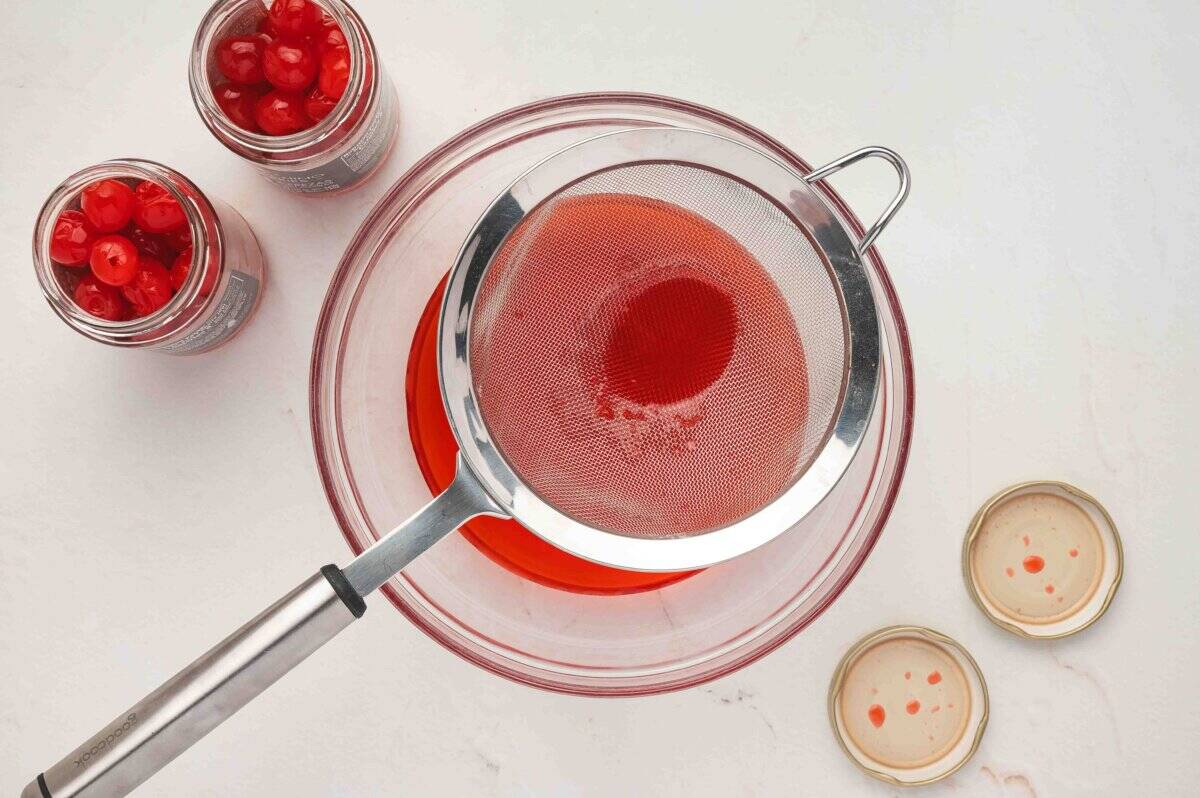 Draining maraschino cherry juice through a fine mesh strainer into a glass bowl with opened jars of cherries nearby.