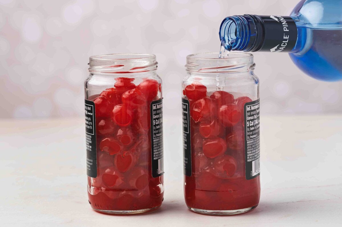 Pouring vodka from a blue bottle into jars filled with drained maraschino cherries.