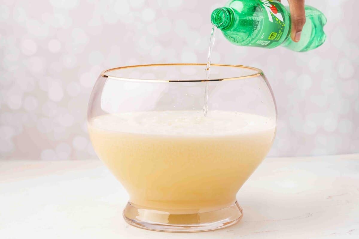 Adding 7UP to the creamy white Christmas punch in a glass punch bowl.