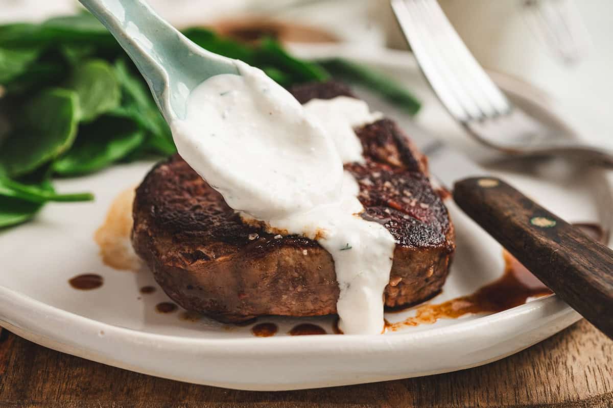 Creamy horseradish sauce being spooned over a cooked steak on a dinner plate.