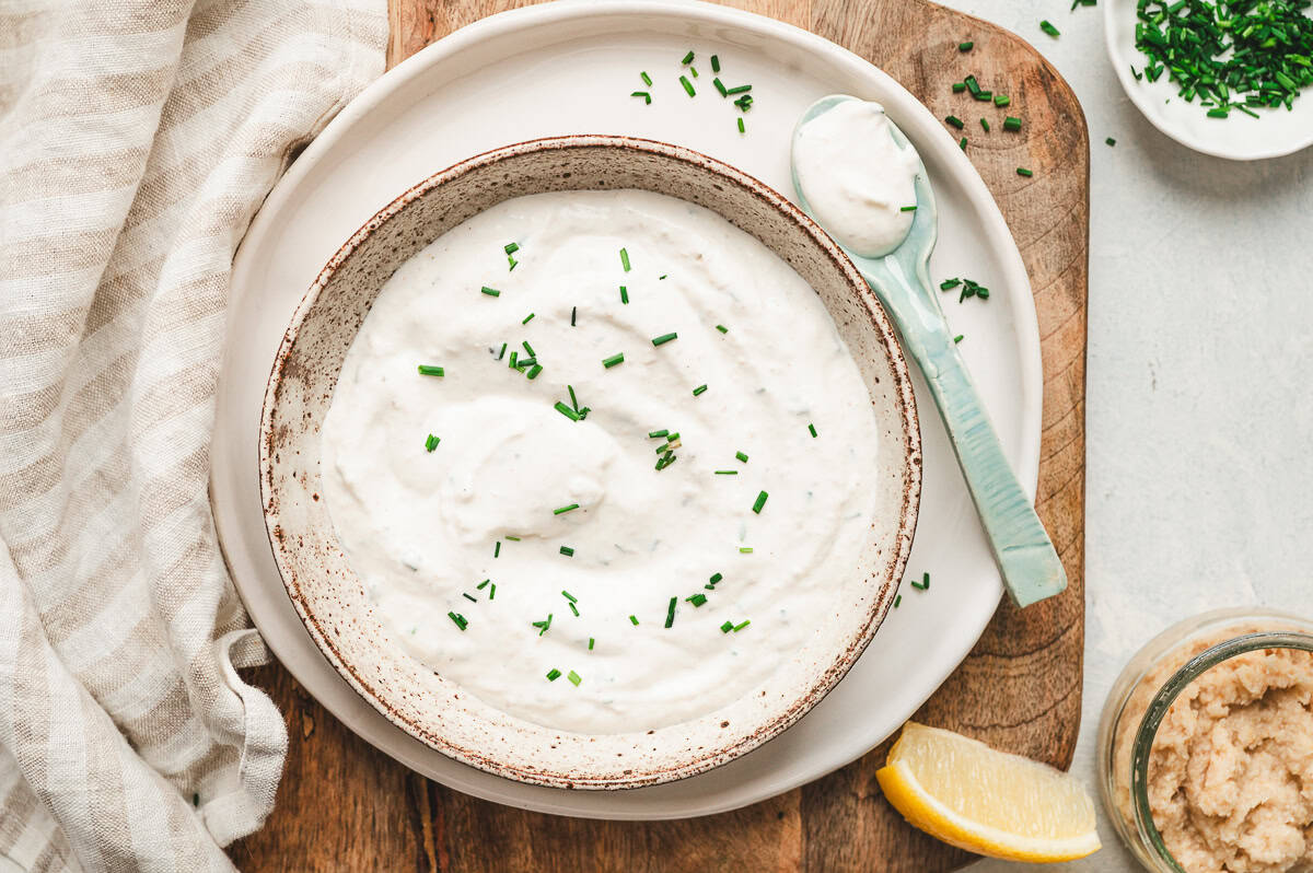 Overhead shot of horseradish sauce in a bowl with chives, a spoon, lemon wedge, and fresh horseradish jar on a wooden board.