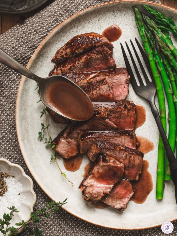 Sliced medium-rare steak on a plate with asparagus, topped with homemade red wine sauce being spooned over the meat.