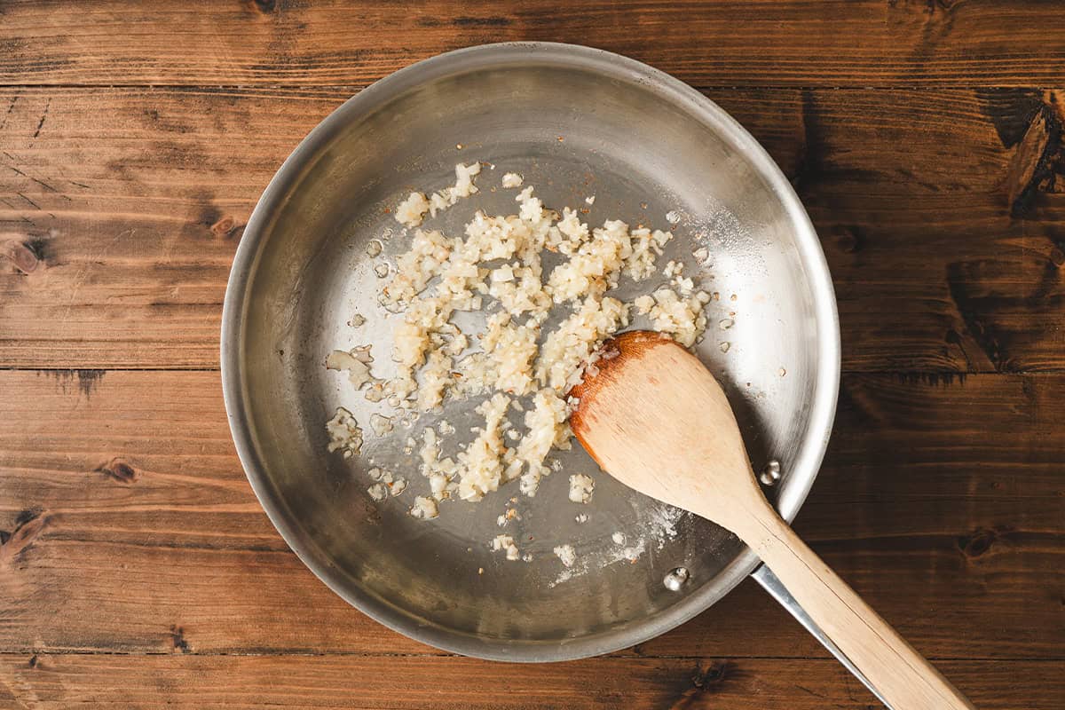 Minced shallots and garlic sautรฉing in a stainless steel pan with a wooden spatula.